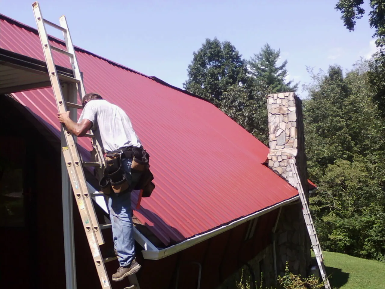 Roofer on a ladder beside a red metal roof and stone chimney — Soffit & Fascia Repair by Roofing Brothers in Battle Ground